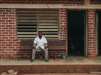 Dr. Sabio sits in white lab coat in front of the brick wall of MSF-supported Bakouma secondary hospital in Central African Republic.