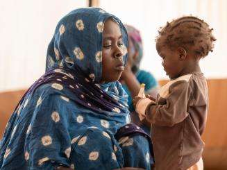 Mother in a blue hijab watches her child eat therapeutic peanut paste in Adré, Chad.